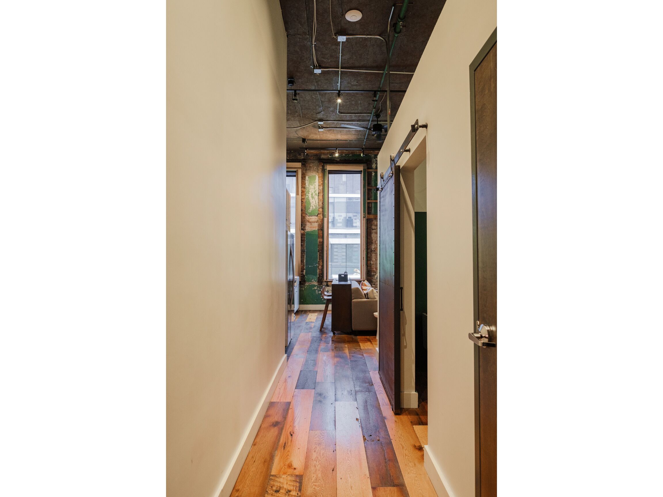 A narrow hallway with wooden flooring, a door on the right, and a window at the end.