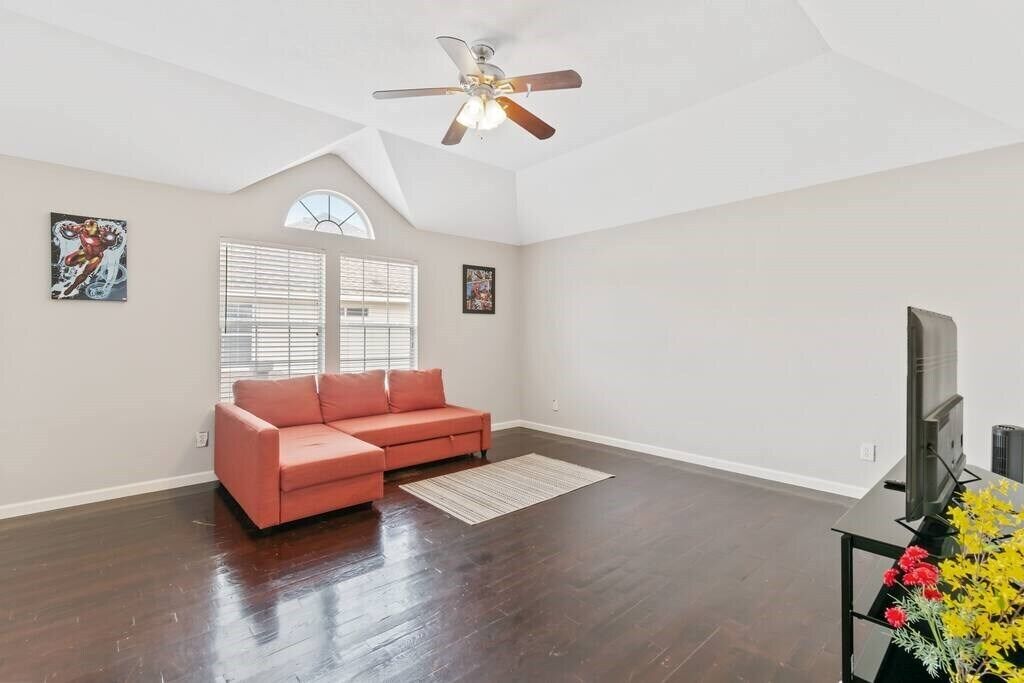 A spacious living room with a dark wood floor, a red sofa, a ceiling fan, and a TV on a stand.