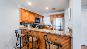 A modern kitchen with wooden cabinets, a granite countertop, and stainless steel appliances. There are three bar stools with leopard print cushions. - Saltwater Dreams @ MP317 | Pools and Beaches (Port Aransas)