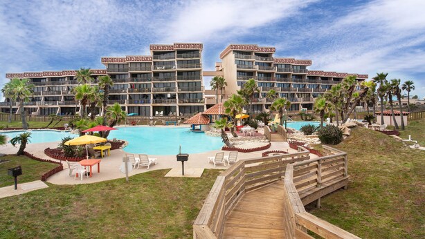 A hotel with a large pool, outdoor seating, and palm trees. - Saltwater Dreams @ MP317 | Pools and Beaches (Port Aransas)