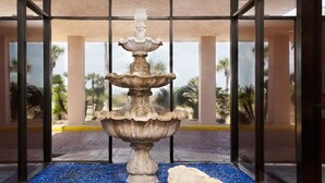A multi-tiered fountain with a stone base, surrounded by blue pebbles, set within a glass enclosure with a view of palm trees and a building in the background. - Saltwater Dreams @ MP317 | Pools and Beaches (Port Aransas)