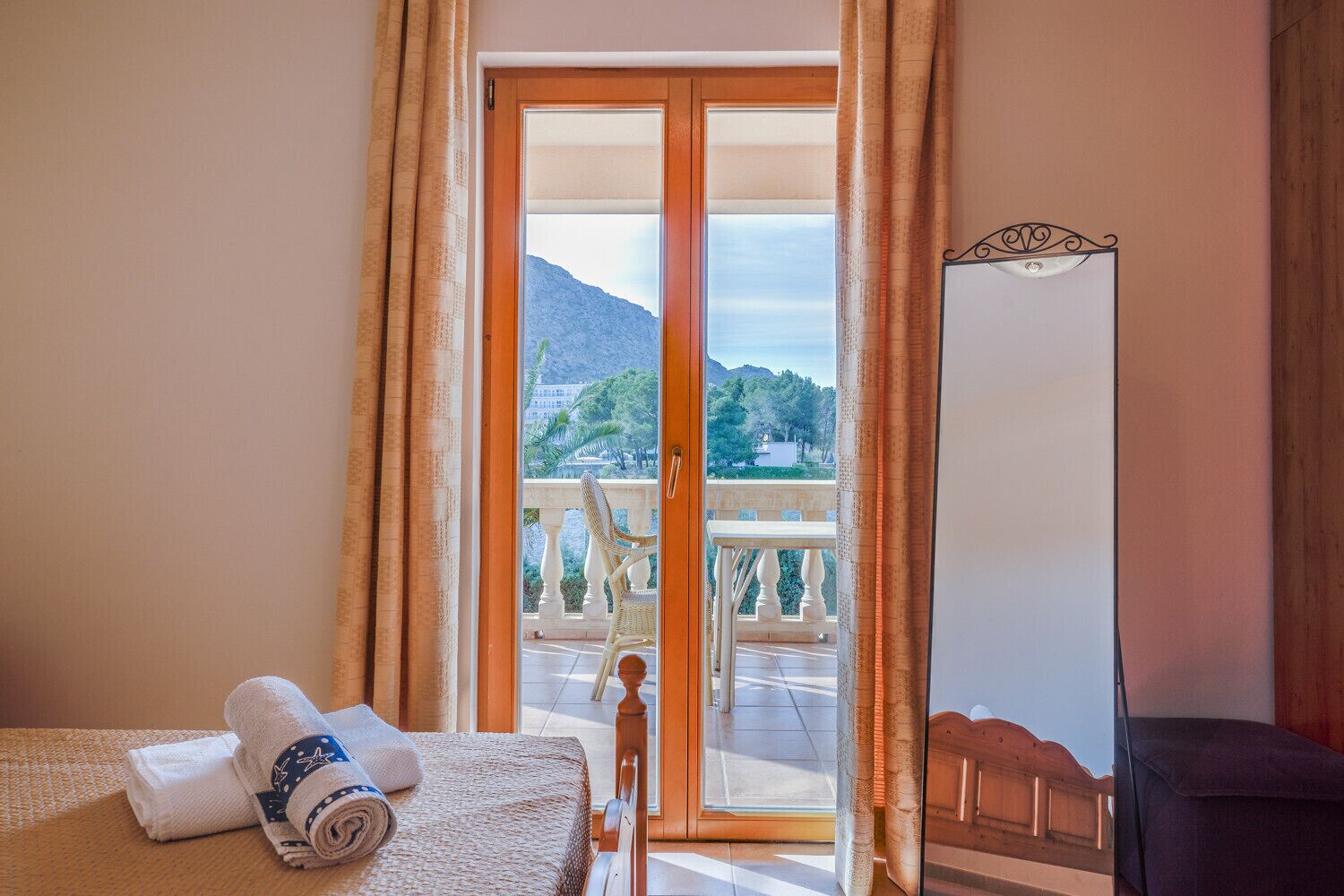 A hotel room with a bed, a chair, a table, and a view of a balcony with a mountain and trees.