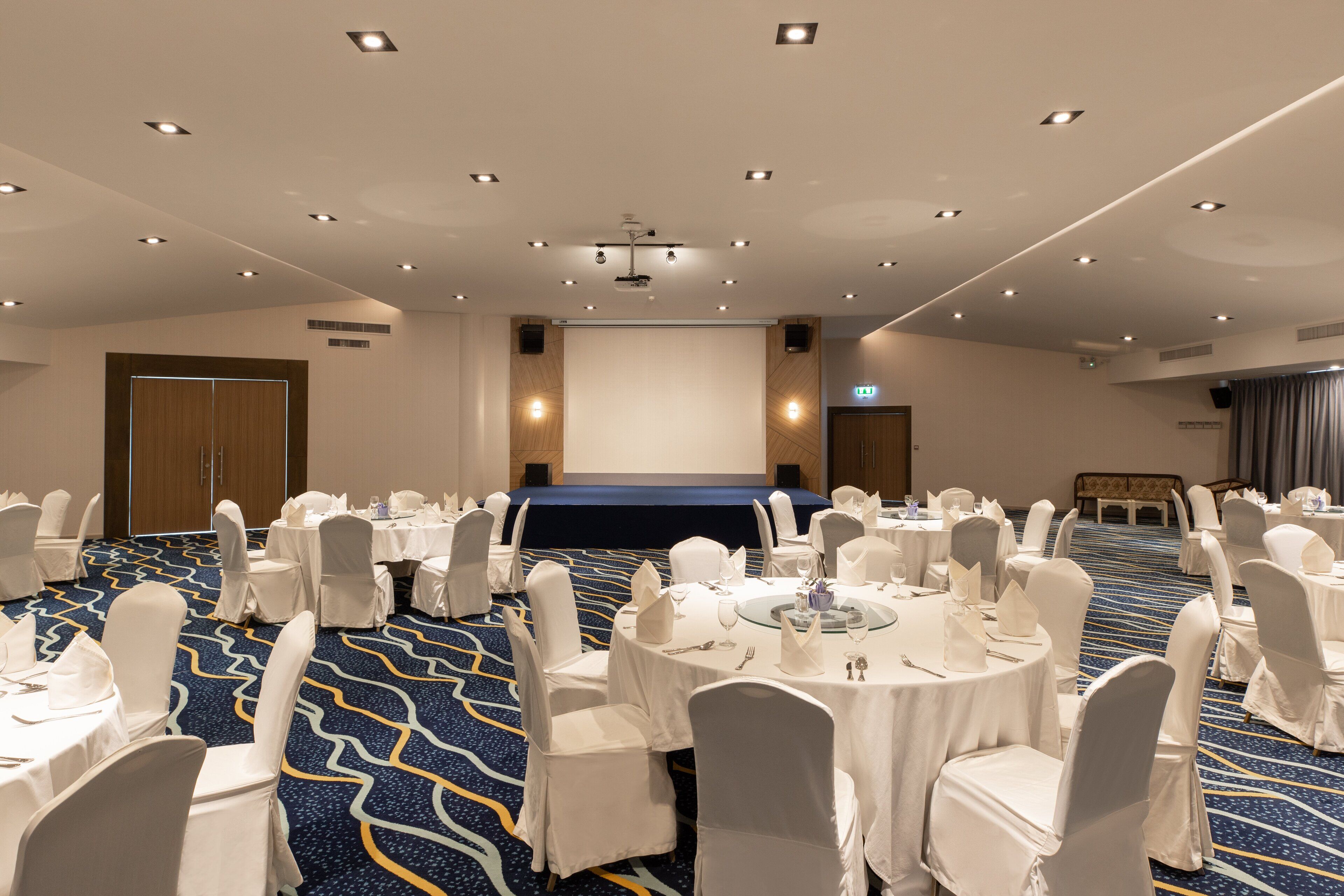 A conference room with round tables, white chairs, and a large screen at the front.