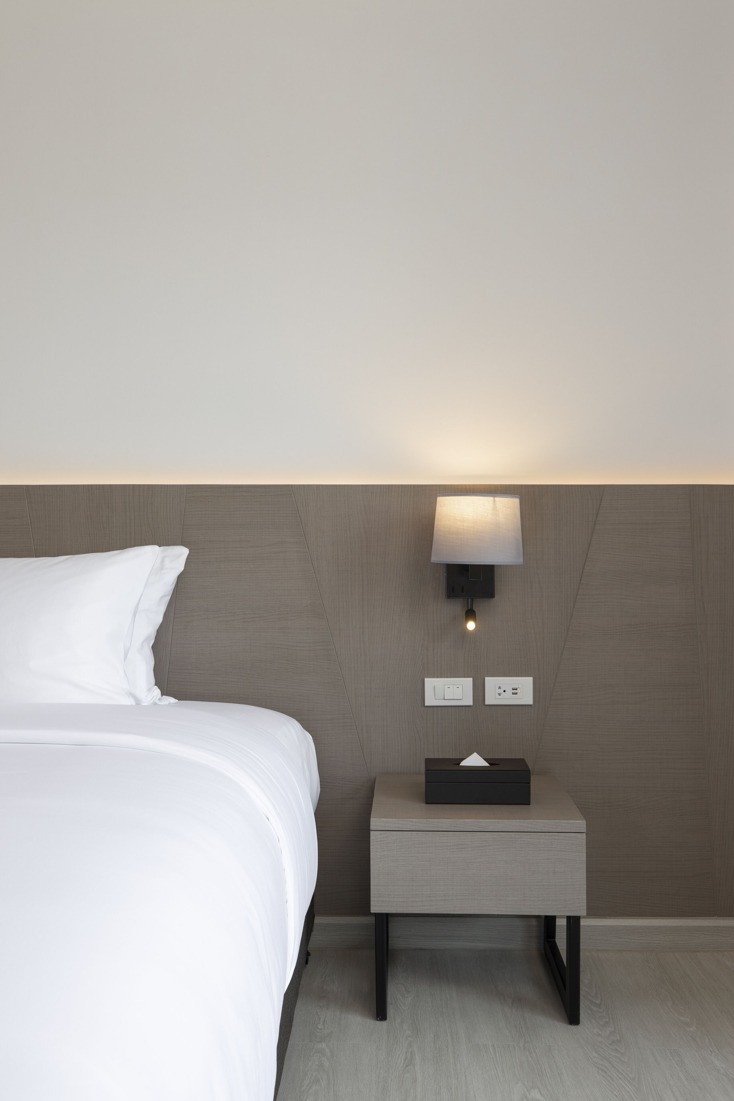 A modern hotel room with a neatly made bed, a bedside table with a tissue box, and a wall-mounted lamp.
