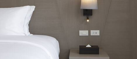 A modern hotel room with a neatly made bed, a bedside table with a tissue box, and a wall-mounted lamp.