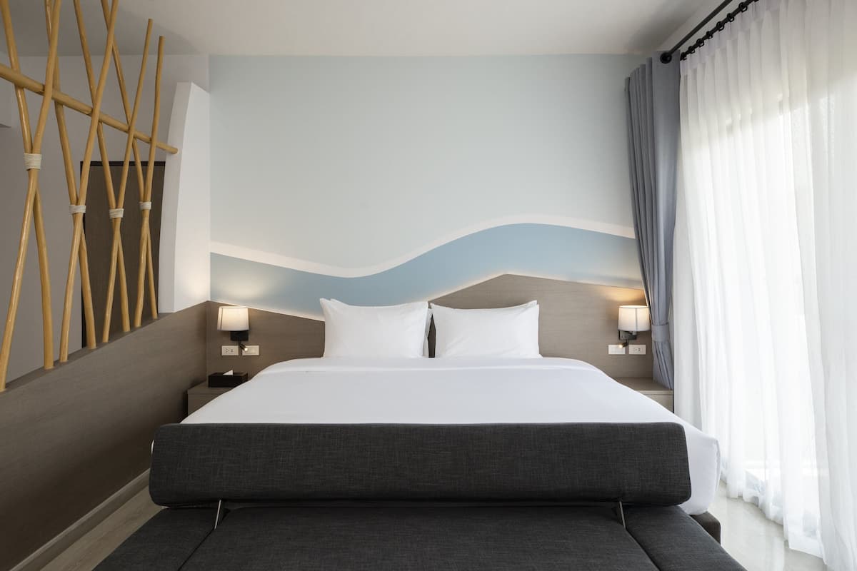 Een moderne hotelkamer met een groot bed, twee bedlampen, een houten wanddecoratie en uitzicht op de omgeving door dunne gordijnen.