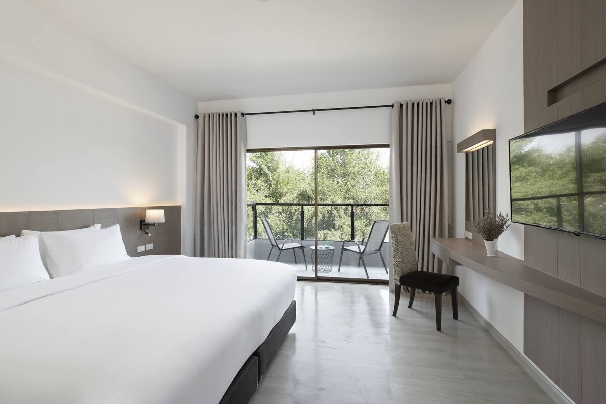A modern hotel room with a large bed, a balcony with a table and chairs, and a TV mounted on the wall.