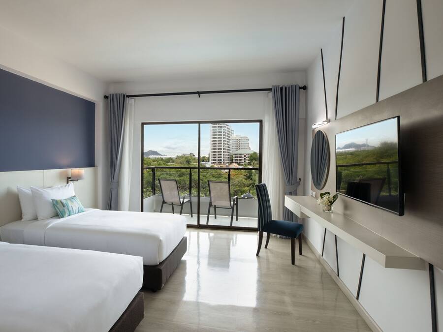 A modern hotel room with two beds, a balcony with a view of a cityscape, and a flat-screen TV mounted on the wall.