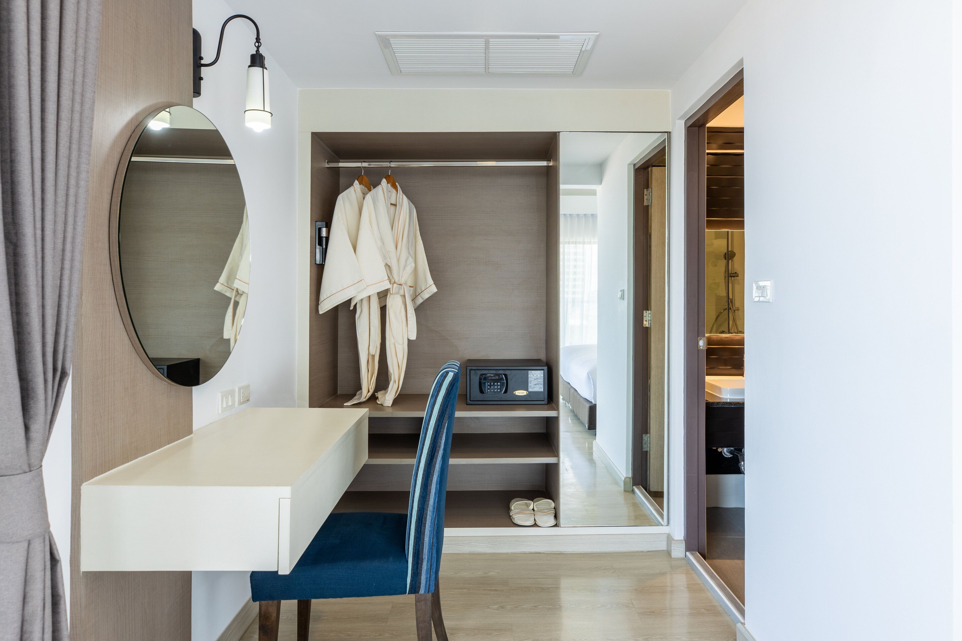 A modern bathroom with a large mirror, a white sink, a blue chair, and a walk-in closet with hanging robes.