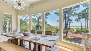 A dining area with a large table, chairs, and a view of the beach and trees through large windows. - Luxurious Ocean Front Home w/ Rec Room (Hilton Head Island)