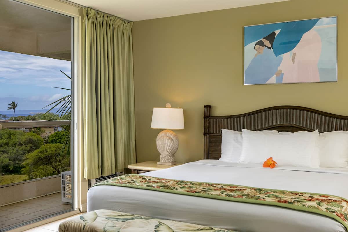 Quarto de hotel com uma cama grande, uma mesa de cabeceira com abajur, um quadro na parede e vista para o verde lá fora.