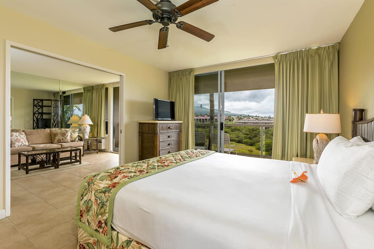 Quarto de hotel com uma cama grande, um ventilador de teto, uma televisão, varanda com vista e um sofá no quarto adjacente.