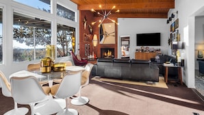 Una sala de estar moderna con chimenea, un comedor con sillas blancas y un ventanal con vistas a árboles.