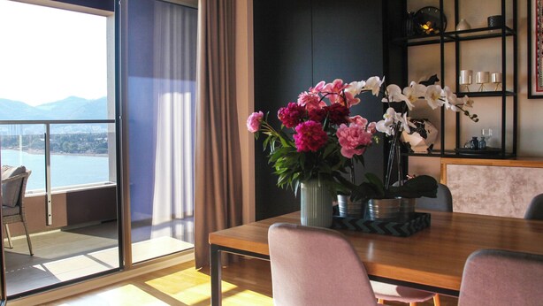 A dining area with a view of a body of water and mountains, a vase with flowers, and a shelf with decorative items. - Sea view Apartment Hermes (Budva)