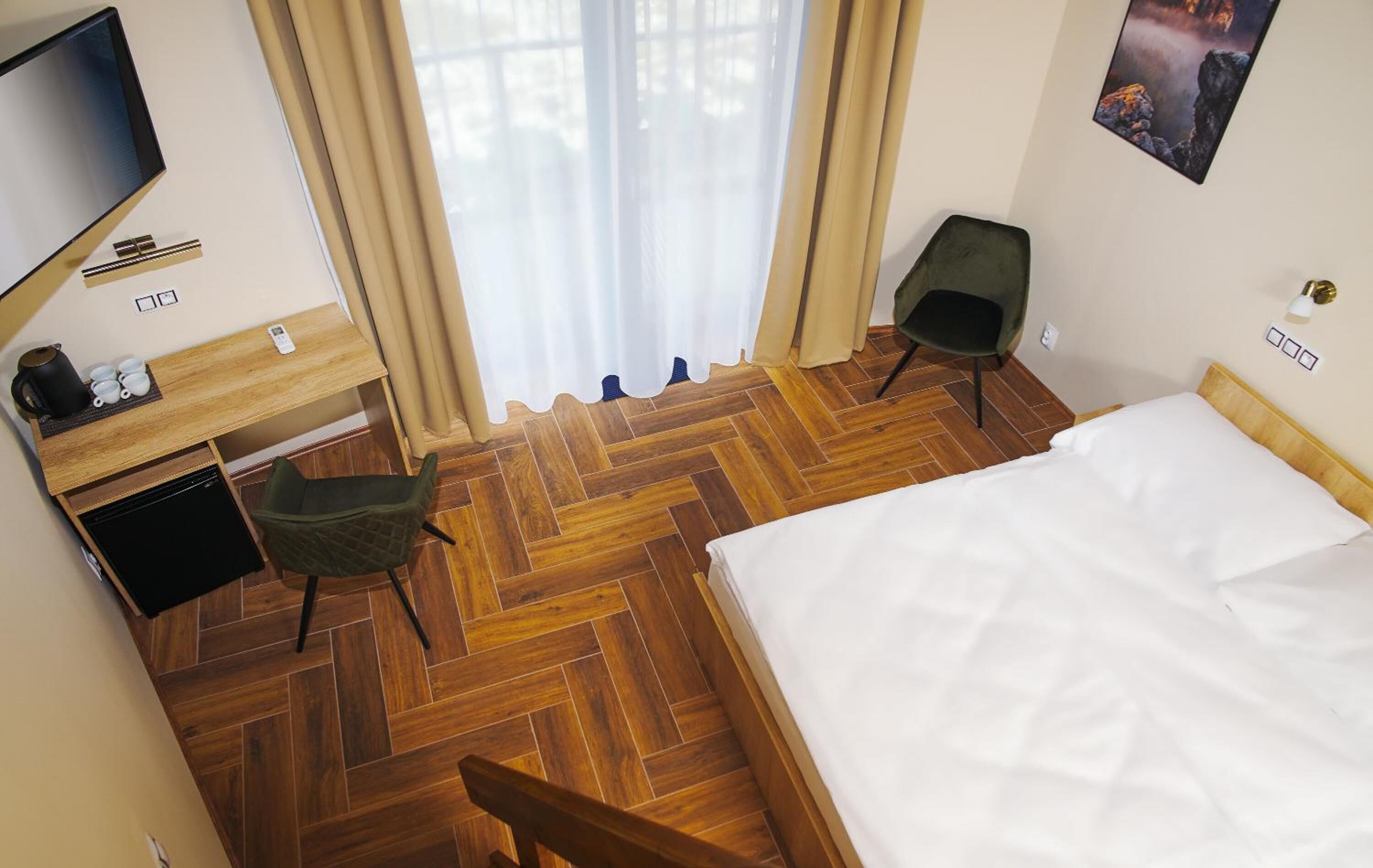 A hotel room with a wooden herringbone floor, a bed with white linens, a desk with a television, a chair, and a window with sheer curtains.
