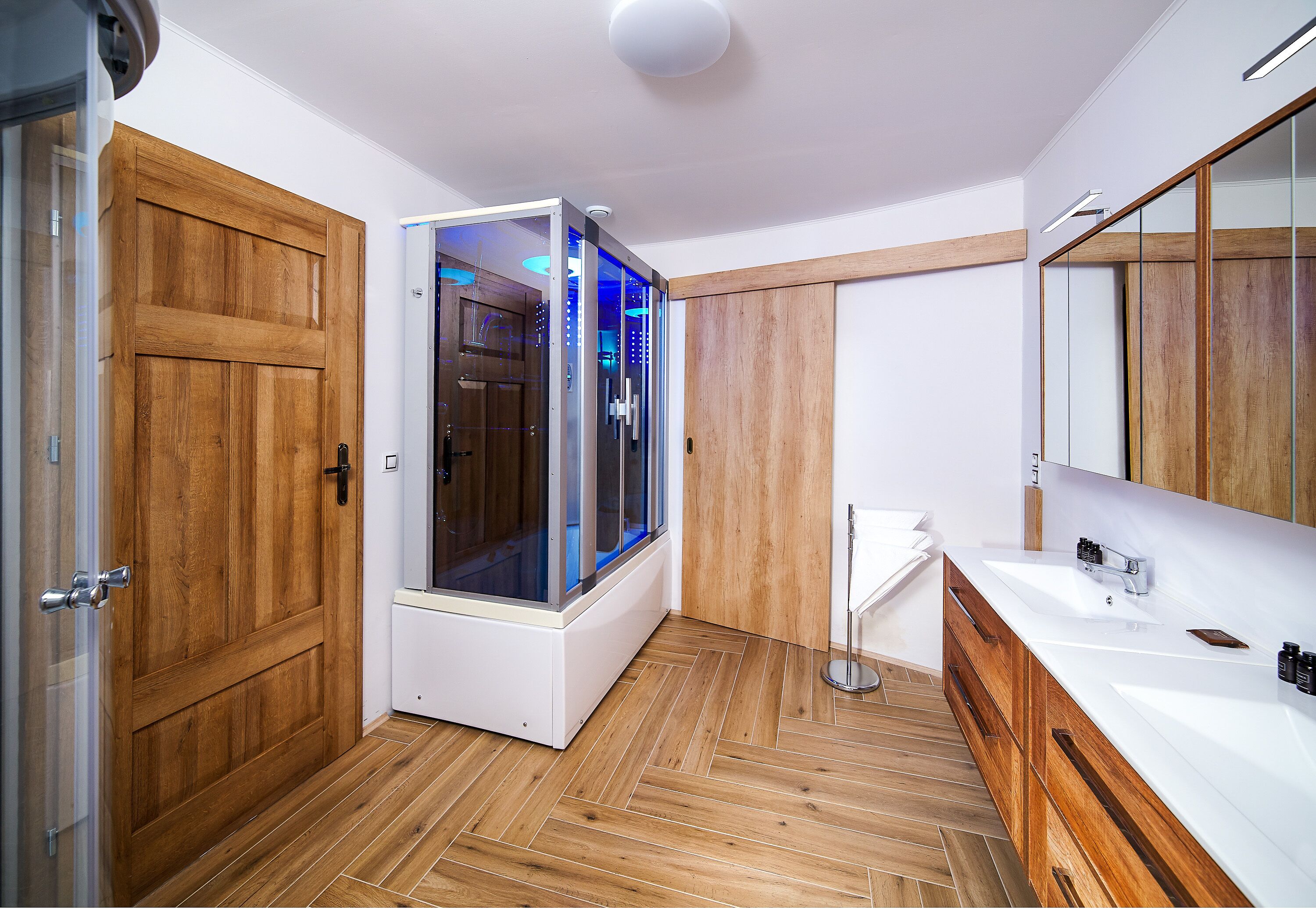 Un baño moderno con suelo de madera, cabina de ducha de cristal, lavabo blanco y tocador de madera.