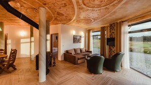 A spacious living area with a wooden ceiling, a sofa, a television, and a large window offering a view of a landscape. - Hotel Dymník (Rumburk)