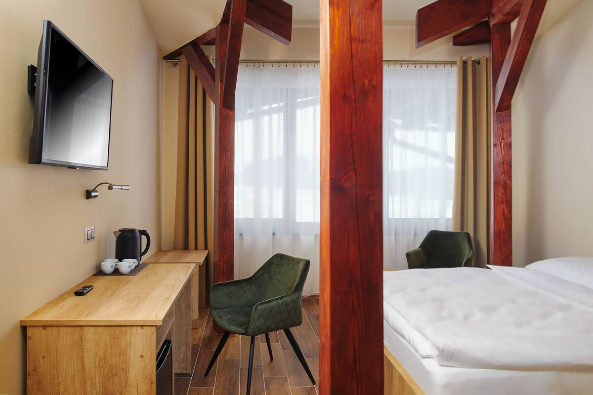 A hotel room with a wooden desk, a green chair, a bed, a TV mounted on the wall, and a window with curtains.
