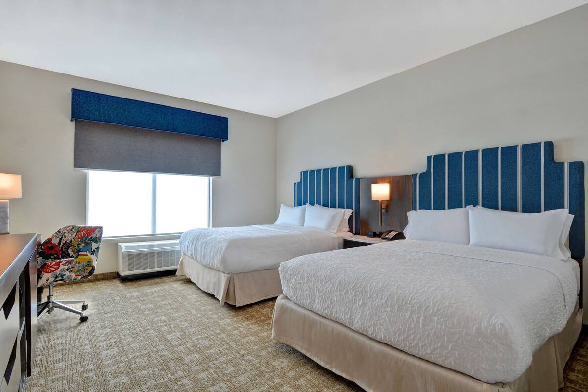 A hotel room with two beds, a desk, a chair, and a large window.
