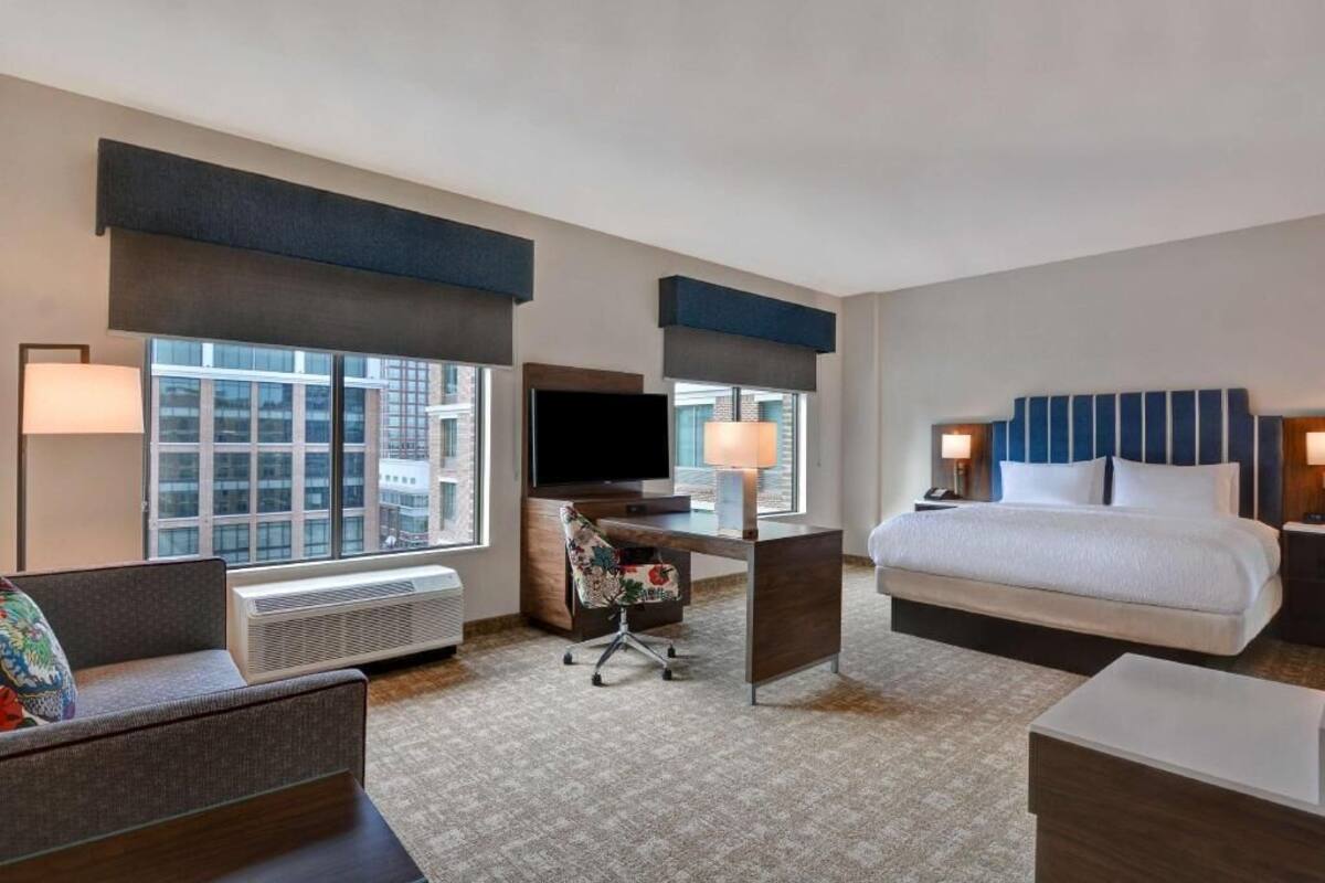 A hotel room with a bed, desk, chair, TV, and large windows.