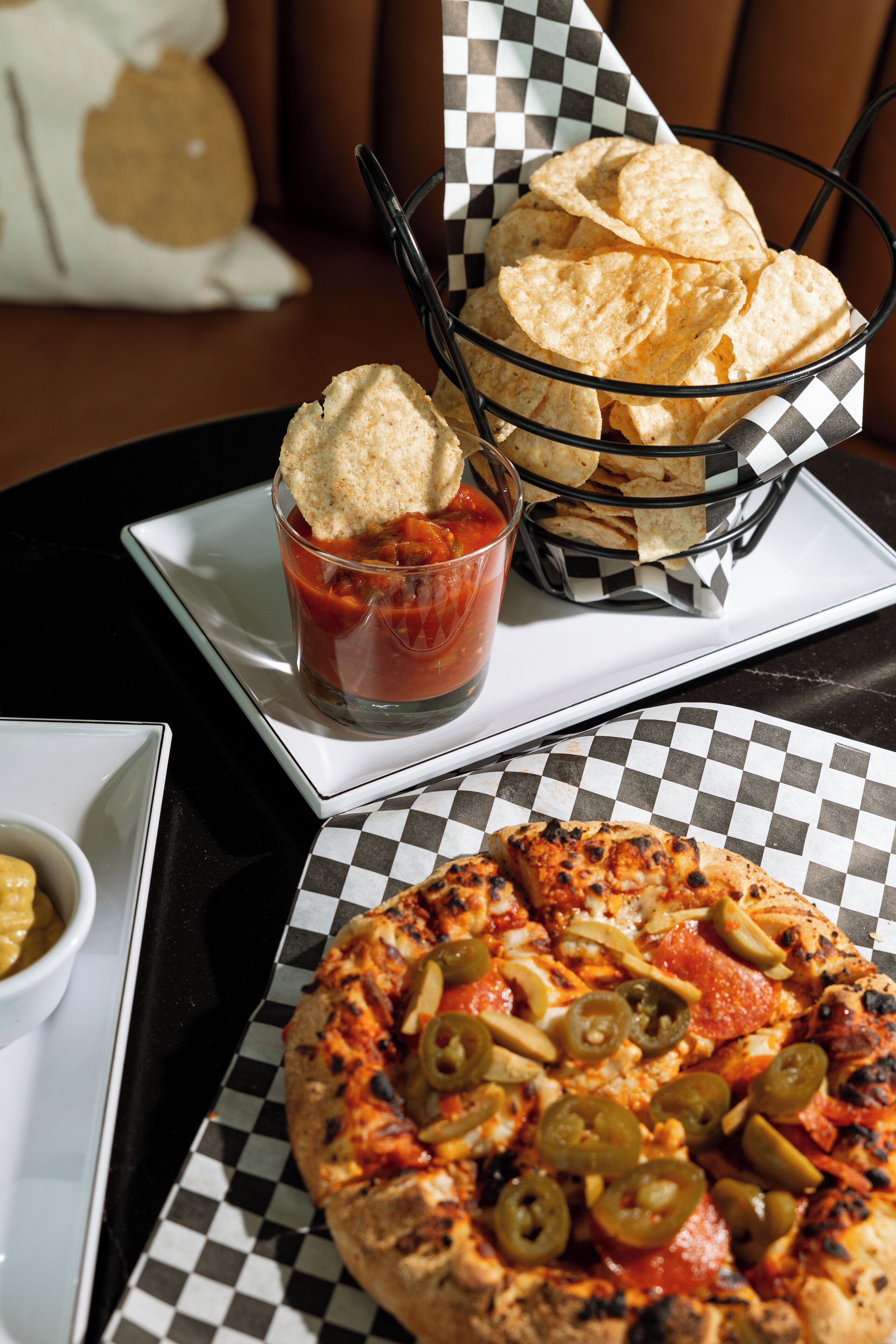 A pizza with jalapeños and a basket of tortilla chips on a checkered tablecloth.