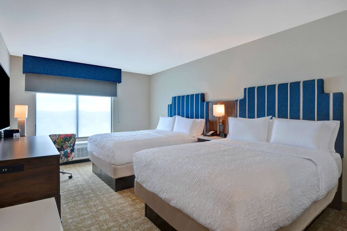 A hotel room with two beds, a desk, a chair, a TV, and a large window with a view.