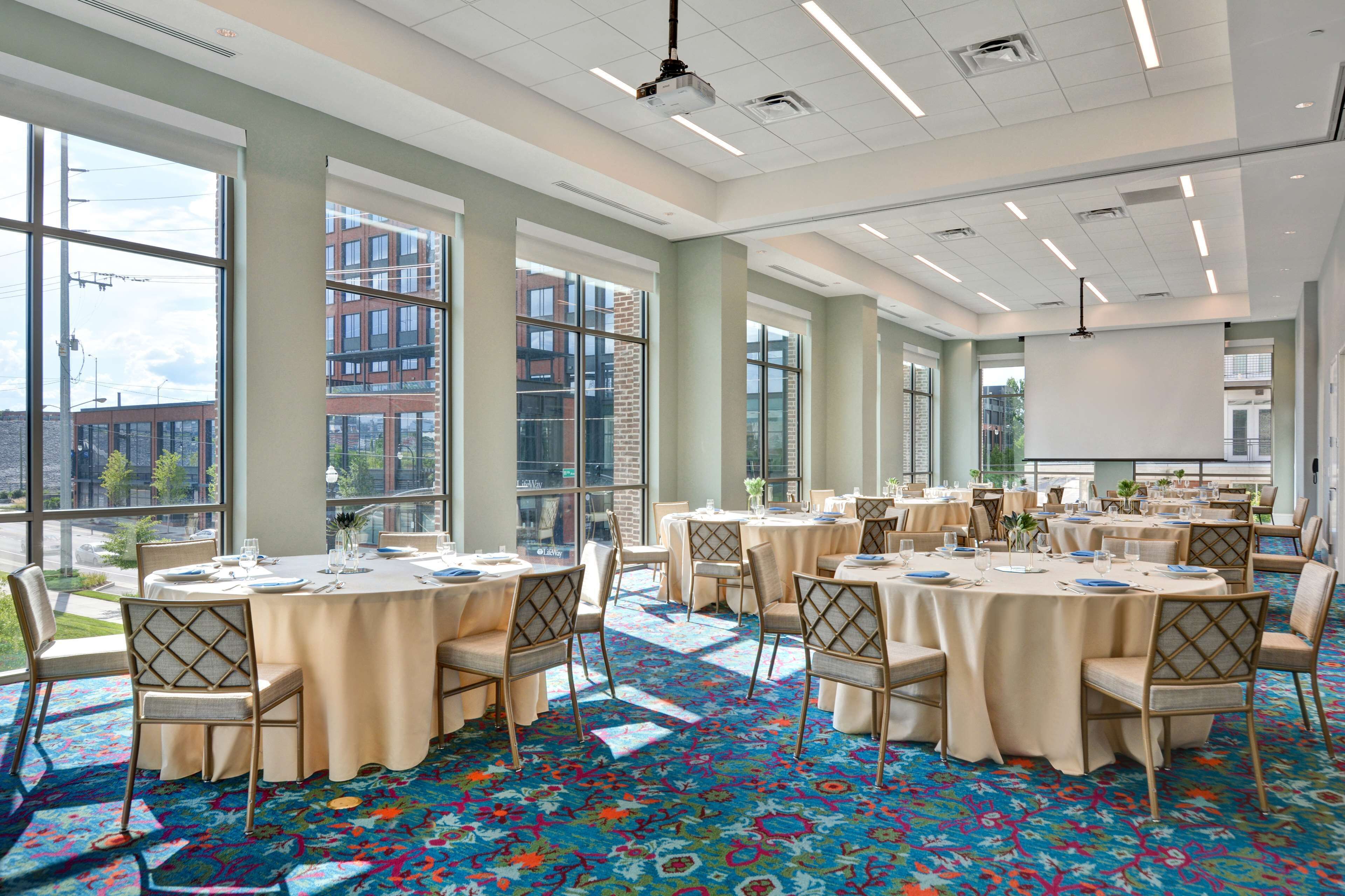A spacious conference room with large windows, round tables, and chairs, featuring a colorful carpet and a projector screen.