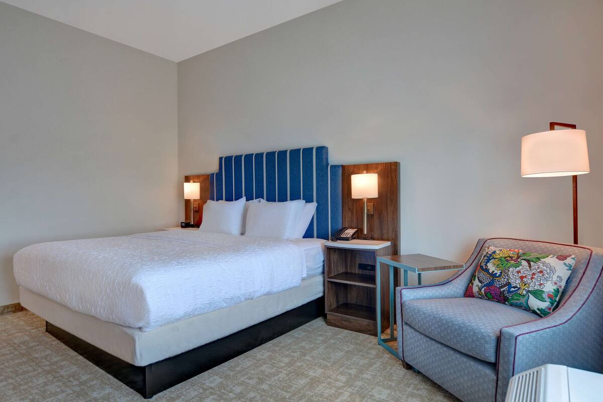 A hotel room with a large bed, bedside tables, a chair, and a lamp.