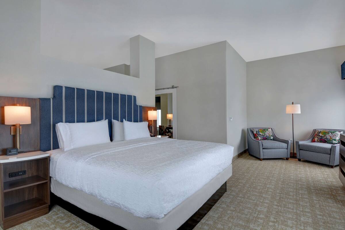 A hotel room with a large bed, two armchairs, a nightstand, and a mirror.