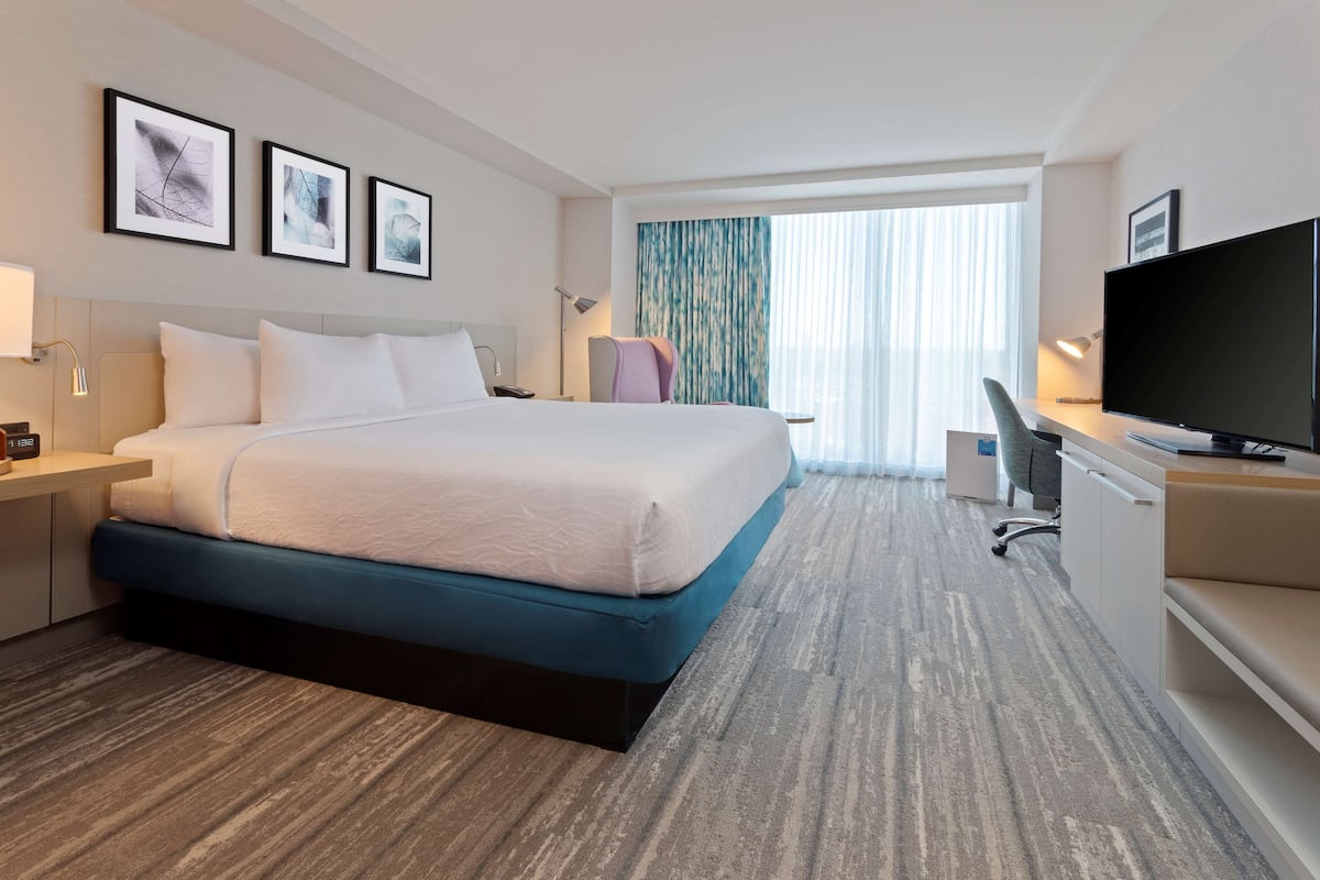 Ein Hotelzimmer mit einem großen Bett, einem Schreibtisch mit Computer, einem Fernseher und gerahmten Kunstwerken an der Wand.