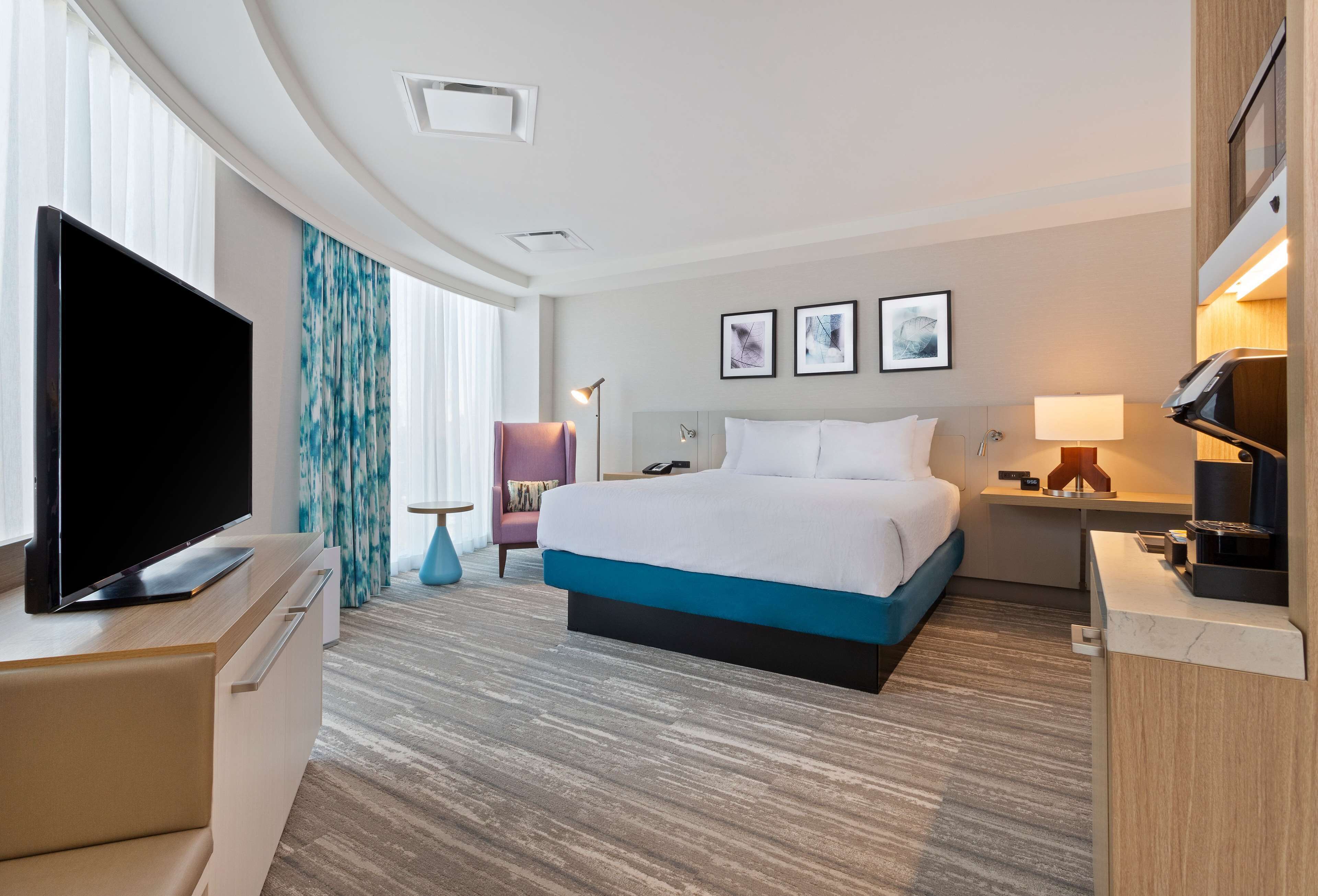 Una habitación de hotel moderna con una cama grande, un televisor de pantalla plana, un escritorio con lámpara y vistas a la ciudad.