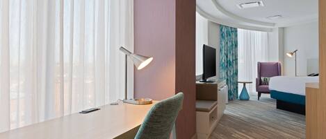 A hotel room with a desk, chair, bed, TV, and a view of the city through large windows.