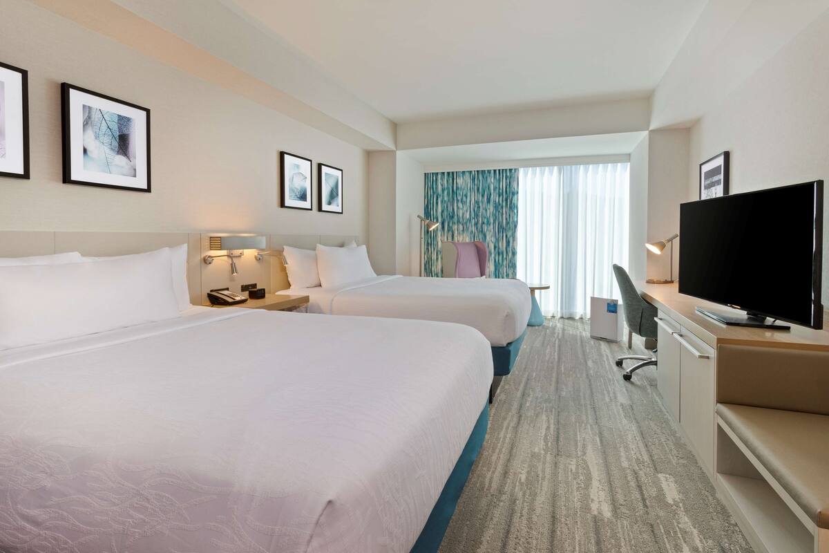 Ein Hotelzimmer mit zwei Betten, einem Schreibtisch, einem Fernseher und einem Fenster mit Vorhängen.