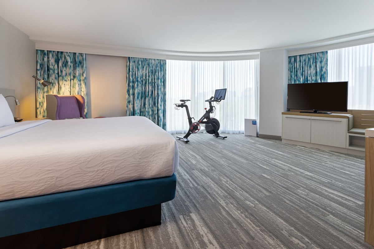 Ein Hotelzimmer mit einem großen Bett, einem Ergometer, einem Flachbildfernseher und einem großen Fenster mit Vorhängen.