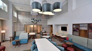 A modern hotel lobby with a long dining table, blue sofas, and red ottomans. There are multiple hanging pendant lights and a large flat-screen TV on the wall. - Hampton Inn & Suites by HIlton Charlotte North I 485 (Charlotte)