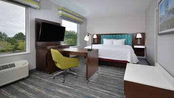 A hotel room with a bed, a desk with a chair, a television, two bedside lamps, and a large window with a view of trees.