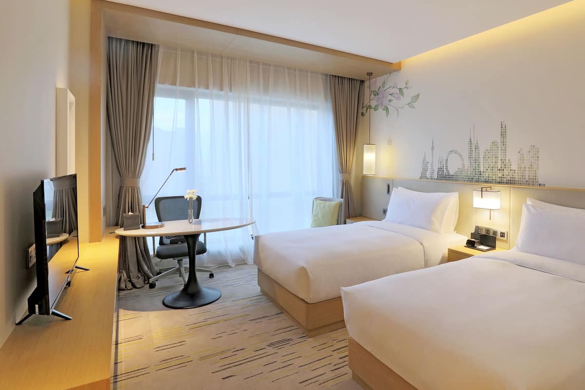 Habitación de hotel con dos camas, un escritorio con silla, televisión y un mural con vista a la ciudad.