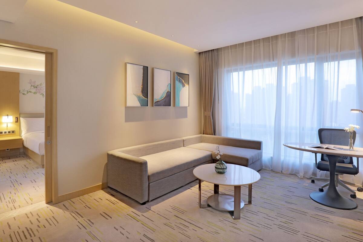 Habitación de hotel con sofá, una mesita redonda, escritorio con silla, lámpara y cuadros en la pared.