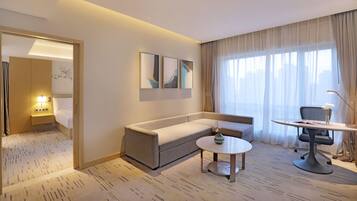 A hotel room with a sofa, a small round table, a desk with a chair, a lamp, and artwork on the wall.