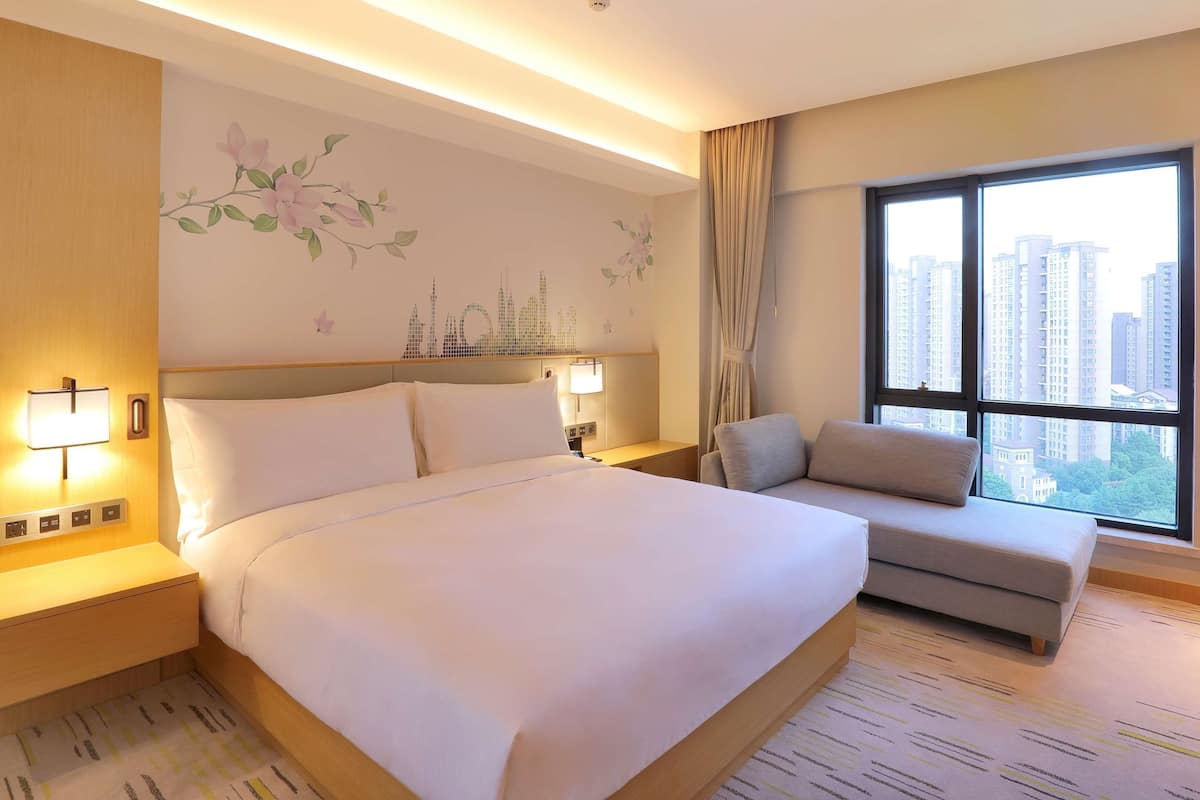 Habitación de hotel con una cama grande, un sofá, una mesita de noche con lámpara, una pared decorada con motivos florales y una ventana con vistas a la ciudad.