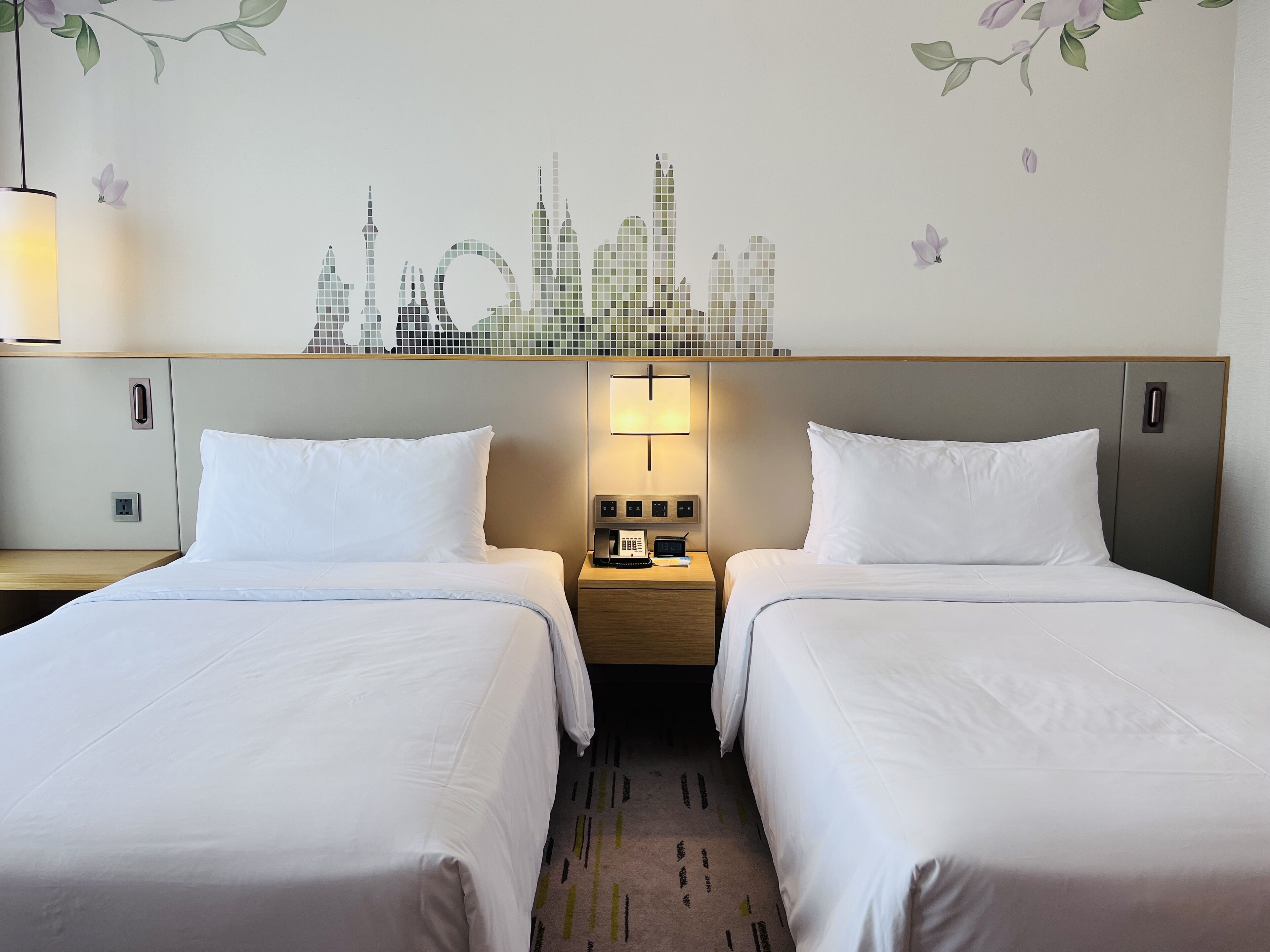 A hotel room with two beds, a cityscape wall mural, a nightstand with a lamp, and a power outlet.