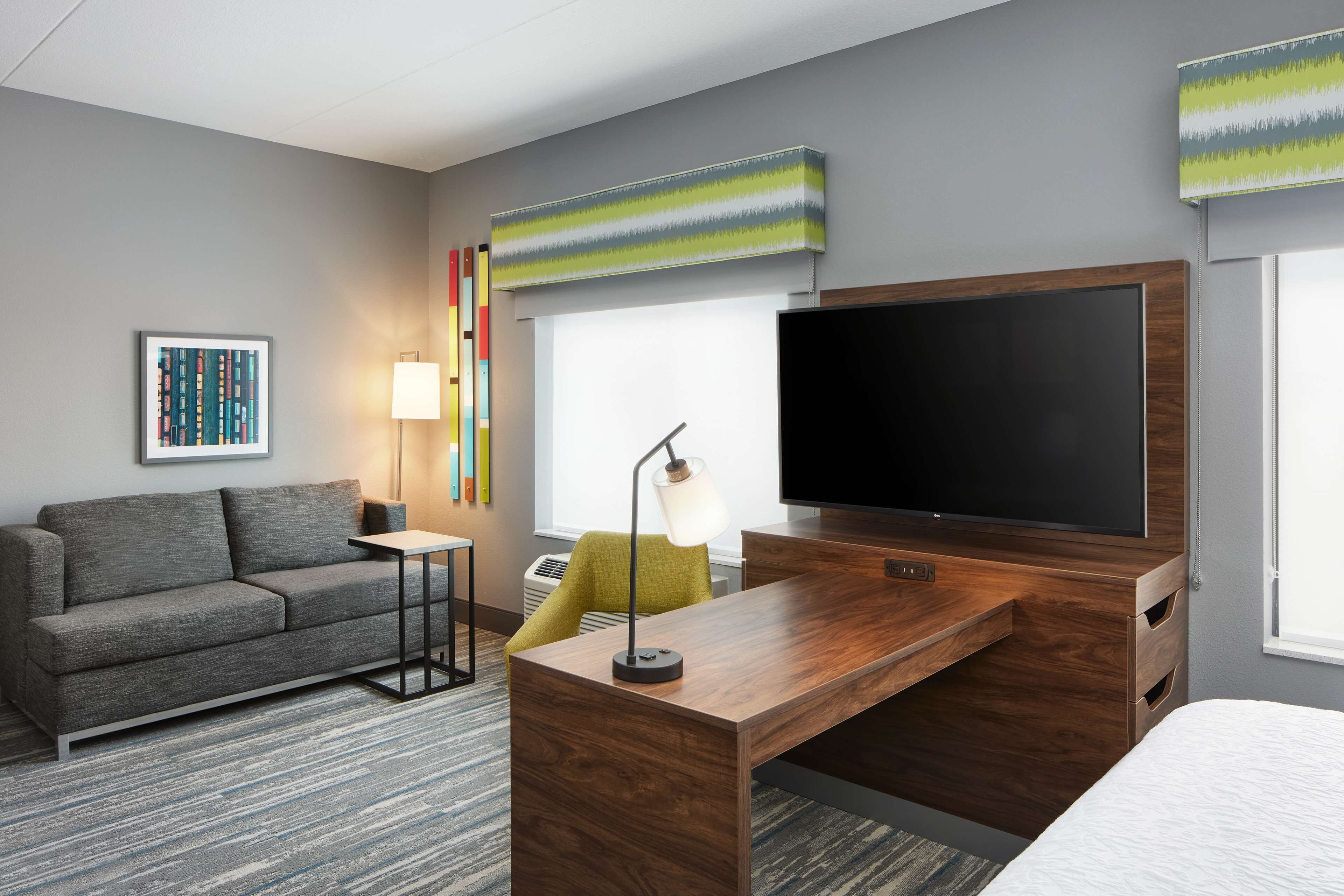 Habitación de hotel con sofá gris, escritorio de madera, televisor de pantalla plana, lámpara y una obra de arte colorida en la pared.