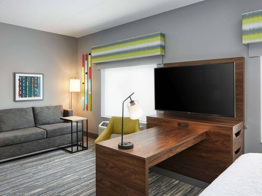 Habitación de hotel con sofá gris, escritorio de madera, televisor de pantalla plana, lámpara y una obra de arte colorida en la pared.