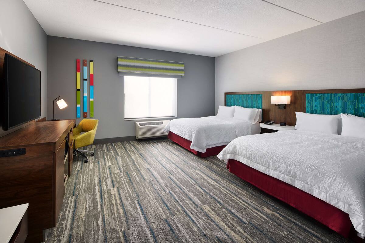 Habitación de hotel con dos camas, un escritorio, un televisor, un ventanal y una decoración de pared colorida.