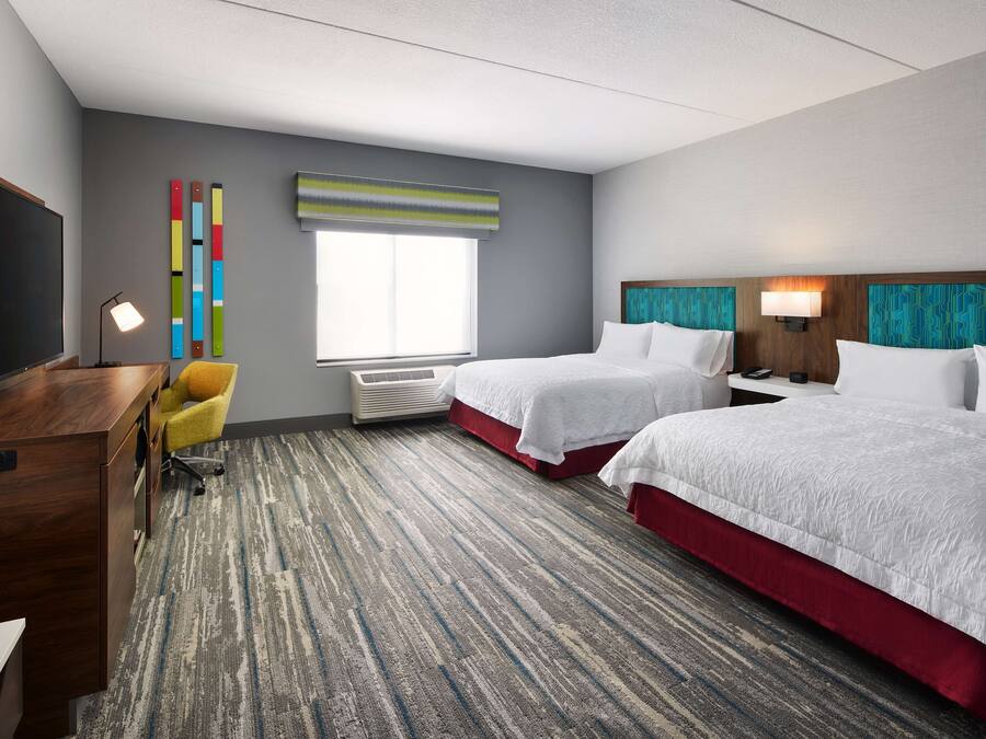 A hotel room with two beds, a desk, a TV, a window, and a colorful wall decoration.