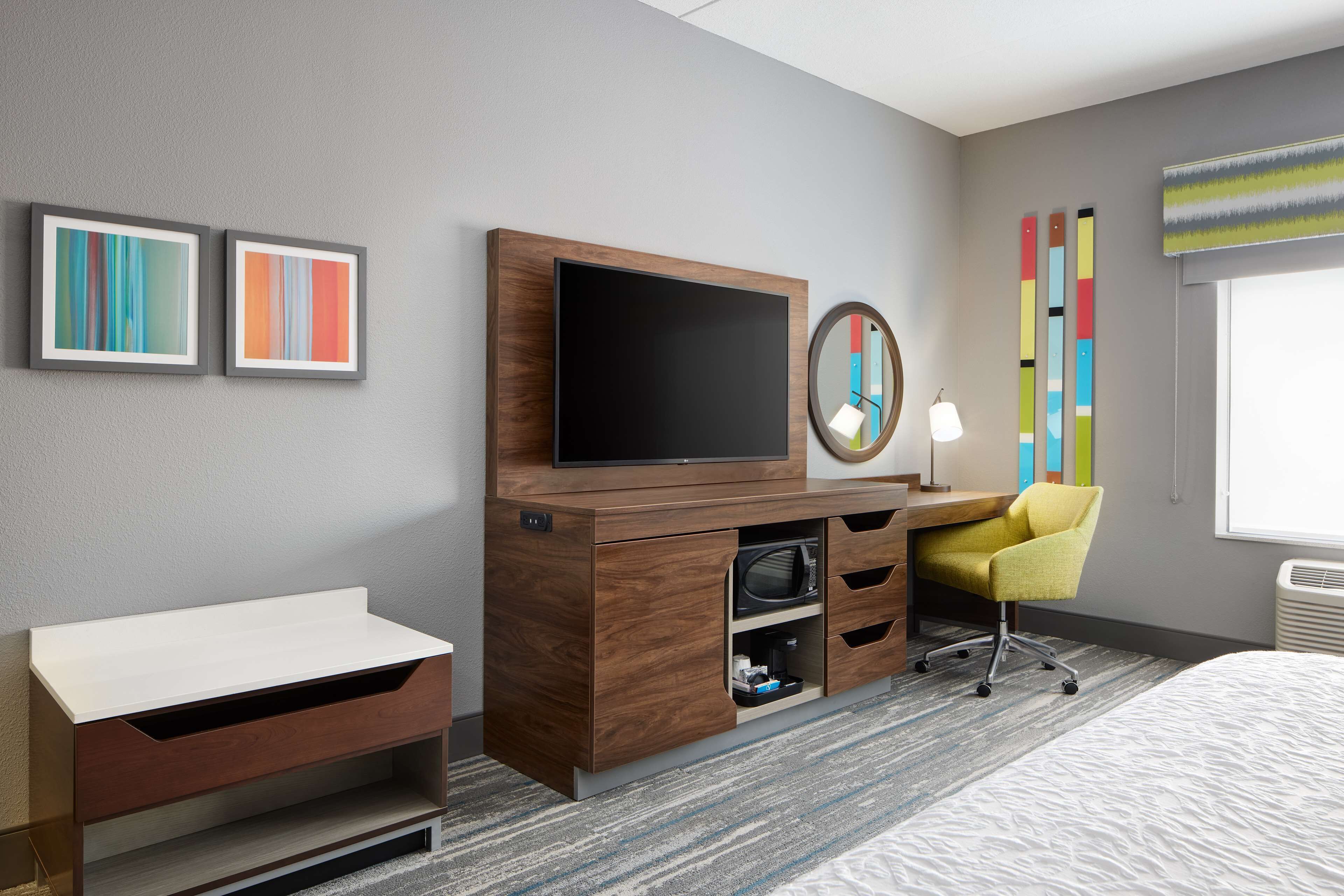 A hotel room with a wooden desk, a flat-screen TV, a yellow chair, a bed, and artwork on the wall.