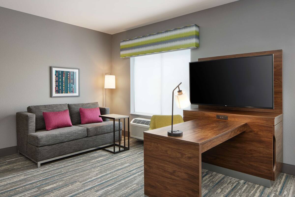 Una habitación de hotel moderna con un sofá gris, un escritorio de madera, un televisor de pantalla plana, un cuadro enmarcado en la pared y un amplio ventanal con vistas.