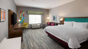 A hotel room with a large bed, a desk with a TV, a chair, a window, and a wall with colorful decor.
