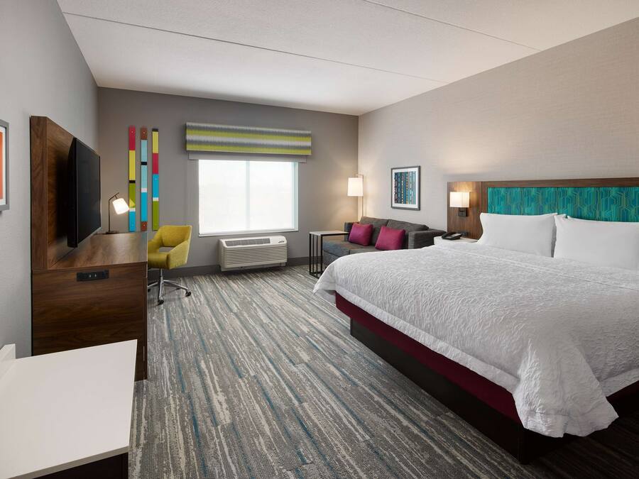 A hotel room with a large bed, a desk with a TV, a chair, a window, and a wall with colorful decor.