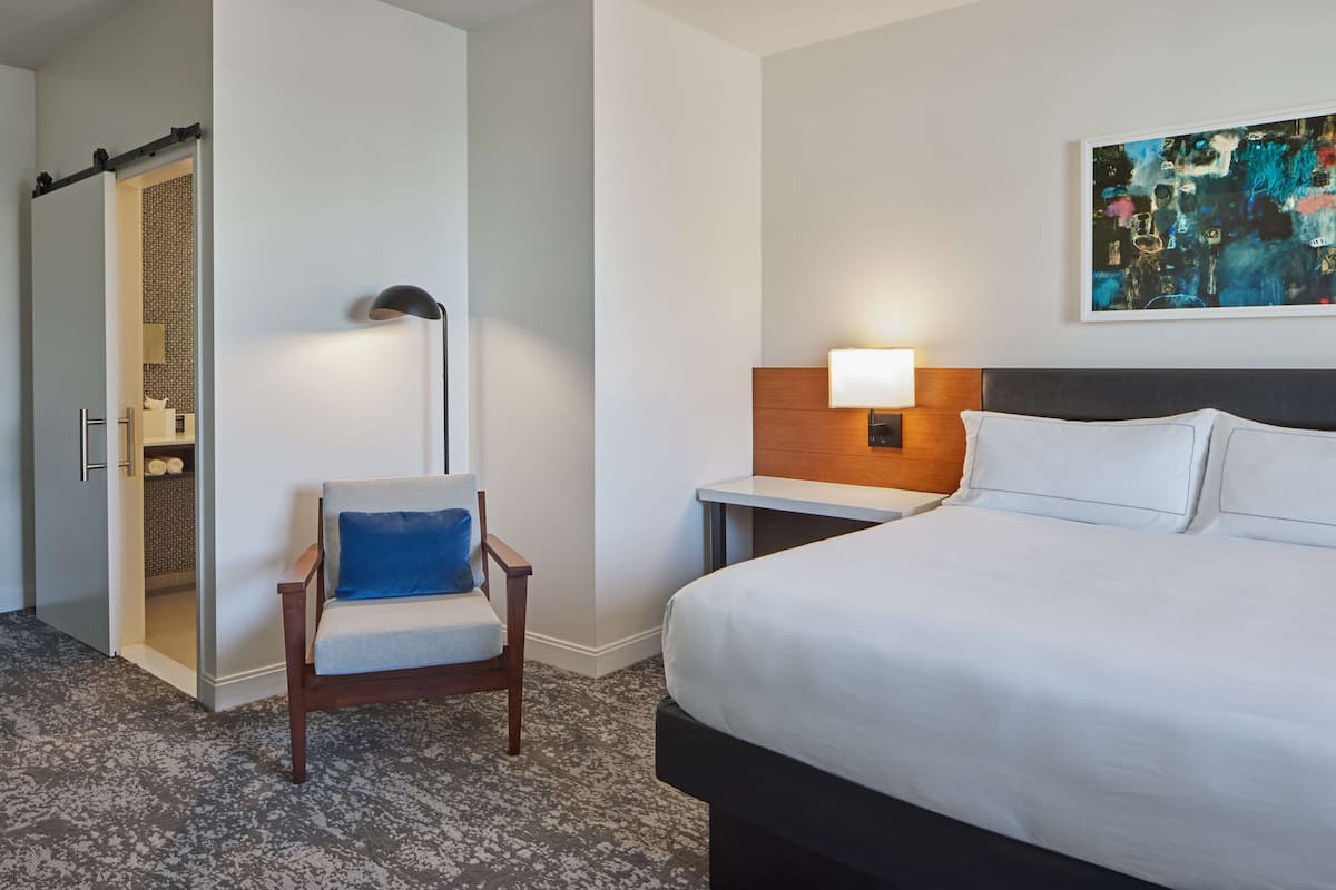Een hotelkamer met een bed, een stoel met een blauw kussen, een nachtkastje met een lamp en een schilderij aan de muur.
